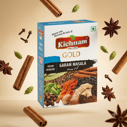 A box of Kichnam Gold Garam Masala spice mix is centered against a warm beige background, surrounded by floating cinnamon sticks, whole spices, and green cardamom pods. The packaging is white and blue with a prominent red Kichnam logo.
