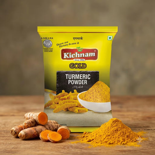 A bag of Kichnam Gold Turmeric Powder standing on a wooden surface, with turmeric roots and loose turmeric powder arranged in front. The package is bright yellow with red and black branding, and the background is a softly blurred warm brown studio setting.